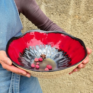Może przedstawiać: Ręcznie robiona ceramiczna miska z żywym czerwonym wnętrzem i czarną obwódką. Misa ma teksturowany, beżowy wygląd zewnętrzny i zawiera małe czerwone jagody i anyż gwiazdkowaty. Miska jest trzymana przez osobę w dżinsowym fartuchu.