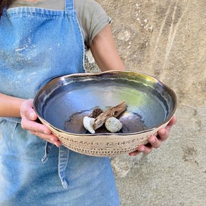 Pode incluir: Uma taça de cerâmica grande, redonda e feita à mão, com um interior de esmalte escuro, metálico e iridescente, e um exterior texturizado de cor clara com padrões geométricos gravados. Dentro da taça há pedaços de madeira e pedras claras.