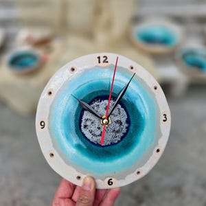 Op de afbeelding: Een ronde keramische wandklok met een verloop van blauwe en turquoise glazuur en een textuurrijk, gespikkeld wit midden. De wijzerplaat heeft zwarte uur- en minutenwijzers, een rode secondewijzer en zwarte cijfers op 12, 3, 6 en 9.