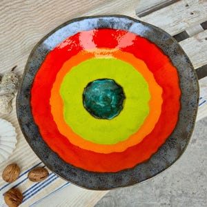 Puede incluir: Un plato de cerámica colorido con anillos concéntricos rojos, naranjas, amarillos y verdes, centrado con un círculo azul turquesa. El plato tiene un borde gris oscuro y se muestra con elementos decorativos como conchas y nueces.