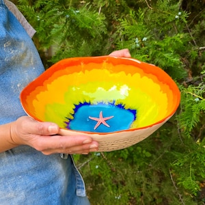 May include: A ceramic bowl with a wavy, uneven rim featuring a vibrant interior gradient of orange, yellow, and lime green, transitioning to a deep blue center. A small, three-dimensional pink starfish is attached to the bottom of the bowl.