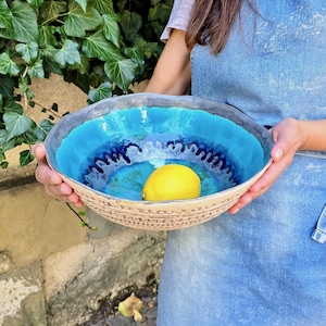 Può includere: Una grande ciotola in ceramica fatta a mano, rotonda, con un esterno testurizzato di colore chiaro caratterizzato da scanalature orizzontali. L'interno è smaltato in vivaci tonalità turchese e blu profondo con un motivo ondulato, contenente un limone.