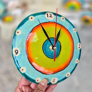 Op de afbeelding: Een ronde keramische wandklok met concentrische ringen in turquoise, oranje en limoengroen. De wijzerplaat heeft zwarte cijfers op de posities 12, 3, 6 en 9, met kleine cirkelvormige markeringen voor de andere uren. De klok heeft zwarte wijzers en een dunne rode secondewijzer.