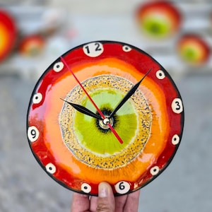 Op de afbeelding: Een ronde wandklok met een levendig, meerkleurig glazuur met concentrische ringen van rood, oranje en geel rondom een fel limoengroen midden. De wijzerplaat bevat zwarte uren- en minutenwijzers, een rode secondewijzer en witte cirkelvormige markeringen met zwarte cijfers op de posities van 12, 3, 6 en 9 uur.