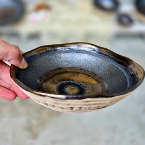 May include: A shallow ceramic bowl with an irregular, wavy rim. The interior features a dark, metallic-toned glaze with a textured, speckled finish and a smooth, golden-brown center. The exterior has a light, matte, textured surface.