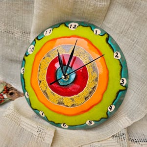 Può includere: Un orologio da parete rotondo con un vivace design a cerchi concentrici multicolore nelle tonalità verde lime, arancione, giallo, rosso e verde acqua. Il quadrante presenta lancette nere e piccole tessere bianche numerate da 1 a 12.