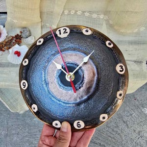 Op de afbeelding: Een ronde keramische wandklok met een donkerblauwe en bronzen reactieve glazuurafwerking. De wijzerplaat toont witte cijfers op de posities 12, 3, 6 en 9, met kleine cirkelvormige markeringen voor de andere uren, witte wijzers en een rode secondewijzer.