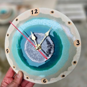 Può includere: Un orologio da parete rotondo in ceramica con una smaltatura sfumata nei toni del crema, turchese e blu scuro. Il quadrante presenta un centro testurizzato, numeri neri a ore 12, 3, 6 e 9, lancette dorate e una sottile lancetta rossa.