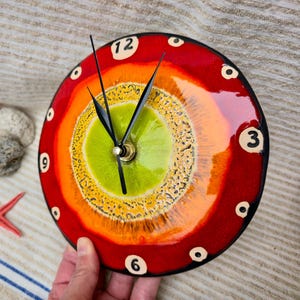 Op de afbeelding: Een ronde, kleurrijke klok met een levendig ontwerp. De wijzerplaat heeft concentrische cirkels van groen, geel, oranje en rood. Zwarte wijzers geven de tijd aan. De klok wordt door een hand vastgehouden. De cijfers 12, 3, 6 en 9 zijn zichtbaar.