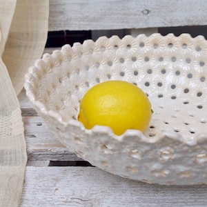 May include: A white ceramic bowl with a lacy design and a single yellow lemon inside. The bowl is sitting on a white wooden surface.