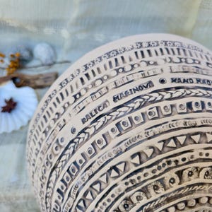 Może przedstawiać: Kremowy, ręcznie robiony ceramiczny wazon ze skomplikowanymi czarnymi geometrycznymi wzorami. Wazon zawiera napis "RALICA MARINOVA HAND MADE" i jest ozdobiony r&oacute;żnymi kształtami i fakturami.