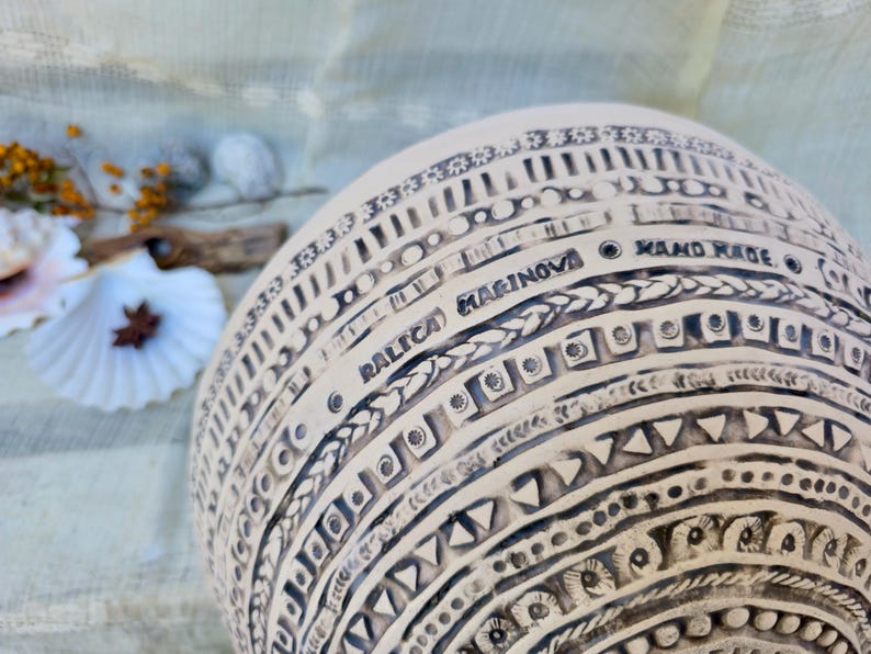 Może przedstawiać: Kremowy, ręcznie robiony ceramiczny wazon ze skomplikowanymi czarnymi geometrycznymi wzorami. Wazon zawiera napis "RALICA MARINOVA HAND MADE" i jest ozdobiony r&oacute;żnymi kształtami i fakturami.