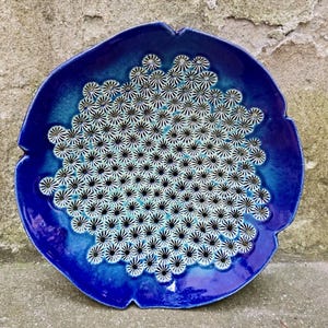 May include: A round ceramic plate with a glossy deep blue glaze featuring a central cluster of small, white, daisy-like flower patterns with black centers. The plate has an irregular, scalloped edge and a smooth, reflective surface finish.