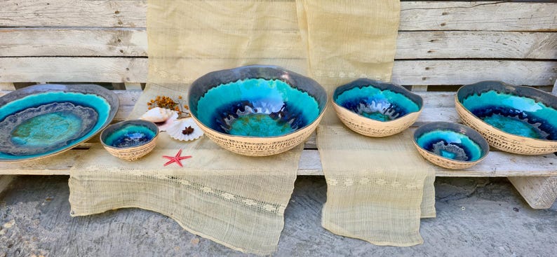 Może przedstawiać: Kolekcja ręcznie robionych ceramicznych misek z żywym turkusowym i niebieskim szkliwem. Miski r&oacute;żnią się rozmiarem i kształtem, z teksturowanym, tkanym wyglądem zewnętrznym. W pobliżu ułożono rozgwiazdę i muszle.