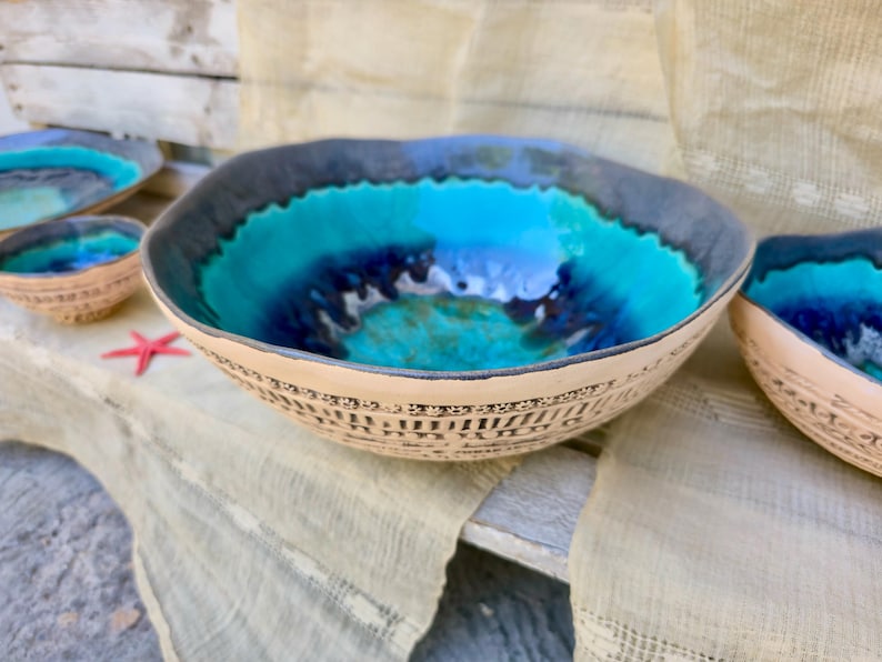 Może przedstawiać: Zestaw ceramicznych misek o morskim designie. Miski mają turkusowe i niebieskie wnętrze, ciemnoszarą obw&oacute;dkę i beżowy zewnętrzny wz&oacute;r. Widoczna jest czerwona rozgwiazda.
