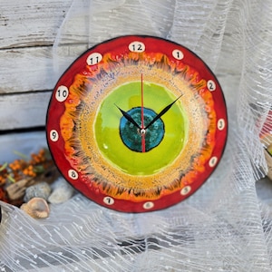 Op de afbeelding: Een ronde keramische wandklok met een levendig, meerkleurig glazuurpatroon dat op een oog lijkt. Het midden is een diepblauwe cirkel omringd door limoengroene, gele en oranje ringen, met een rode buitenrand. De cijfers 1-12 zijn in het zwart.