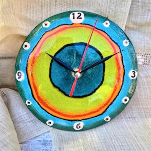 May include: A round, colorful wall clock with a vibrant, abstract design. The clock face features concentric rings of teal, green, orange, and black. Black hands and a red second hand indicate the time. White numbers mark the hours, and small white dots encircle the face.