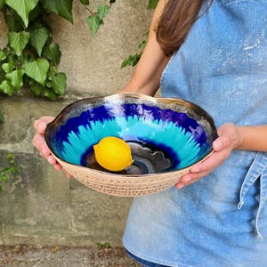 Puede incluir: Un cuenco de cerámica redondo con borde ondulado, que presenta un interior brillante con un degradado de azul profundo y turquesa que pasa a negro. El exterior tiene un acabado texturizado en beige claro. Un limón amarillo brillante descansa dentro.
