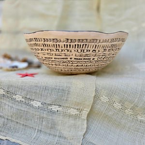 Może przedstawiać: Beżowa ceramiczna miska z falistym brzegiem i skomplikowanymi czarnymi wzorami wytrawionymi. Misa ma poziome pasma z geometrycznymi i tekstowymi wzorami. Widoczne są słowa "Hand Made". Misa stoi na kremowej tkaninie.