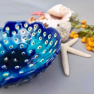 Puede incluir: Un cuenco de cerámica hecho a mano con un vibrante esmalte azul y turquesa. El cuenco tiene una forma orgánica con numerosas pequeñas aberturas circulares. Conchas marinas, una estrella de mar y ramitas de bayas.