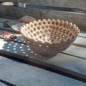 May include: A beige ceramic bowl with a floral pattern and scalloped edges. The bowl is placed on a weathered wooden surface, with a starfish and seashells nearby. The bowl's design features small, star-shaped cutouts.