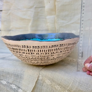 Może przedstawiać: Beżowa ceramiczna miska ze skomplikowanymi czarnymi wzorami i turkusowym wnętrzem. Misa ma ciemnoniebieską obw&oacute;dkę i średnicę około 20 cm. Misa stoi na kremowej tkaninie.