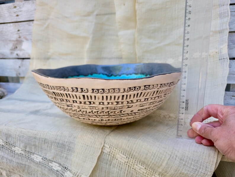Może przedstawiać: Beżowa ceramiczna miska ze skomplikowanymi czarnymi wzorami i turkusowym wnętrzem. Misa ma ciemnoniebieską obw&oacute;dkę i średnicę około 20 cm. Misa stoi na kremowej tkaninie.