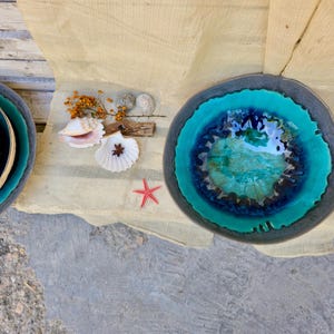 Może przedstawiać: Zestaw ceramicznych misek z turkusowym i ciemnoniebieskim szkliwem. Miski mają unikalny, organiczny kształt i są ułożone na jasnej powierzchni. Obecne są r&oacute;wnież dekoracyjne muszle, rozgwiazda i suszone kwiaty.