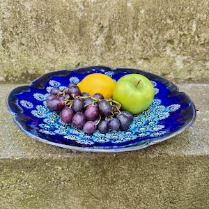 May include: A scalloped ceramic plate featuring a deep blue glaze with a repeating white and light blue floral pattern. The plate holds a cluster of dark purple grapes, a yellow lemon, and a bright green apple, showcasing a vibrant contrast.
