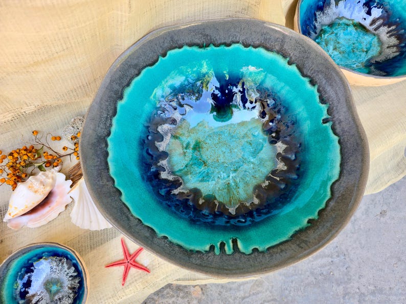 Może przedstawiać: Ceramiczna miska o unikalnym designie. Miska ma żywe turkusowe wnętrze, ciemnoniebieski i brązowy środek oraz szarą obw&oacute;dkę. Muszle, czerwona rozgwiazda i inne elementy dekoracyjne otaczają miskę.