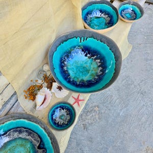 Może przedstawiać: Kolekcja ceramicznych misek z żywym turkusowym i niebieskim szkliwem. Miski r&oacute;żnią się rozmiarem i kształtem, z ciemnoszarą obw&oacute;dką. Niekt&oacute;re miski mają tkaną fakturę. Muszle i rozgwiazda są ułożone w pobliżu.