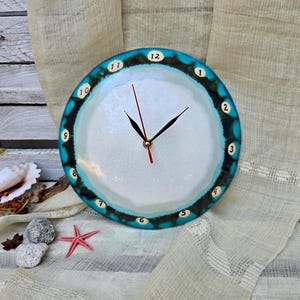 May include: A round wall clock with a white center and a wide, textured border featuring shades of turquoise, blue, and black. The clock face displays numbers 1 through 12 in small white circles along the edge, with black hour and minute hands and a red second hand.