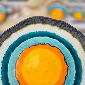 May include: A decorative ceramic plate with a unique design. The plate features concentric circles of color, including yellow, orange, turquoise, white, and a dark gray rim. The plate's irregular edge adds to its artistic appeal.