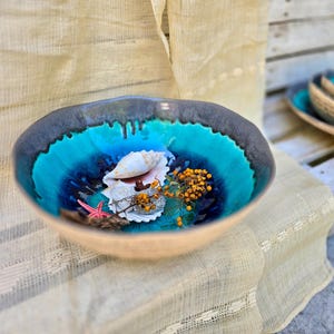 Może przedstawiać: Ceramiczna miska z turkusowym i ciemnoniebieskim szkliwionym wnętrzem, wypełniona muszlami, małą rozgwiazdą i suszonymi ż&oacute;łtymi jagodami. Misa ma beżowy wygląd zewnętrzny i jest prezentowana na kremowej tkaninie.