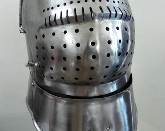 May include: A polished, silver-colored medieval helmet with a rounded top and a face guard. The helmet features numerous small holes for ventilation and a neck guard. It is displayed on a wooden stand with a black strap.