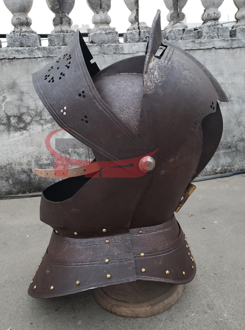 Puede incluir: Un casco medieval con visera y protecci&oacute;n para el cuello. El casco est&aacute; hecho de metal y tiene un acabado marr&oacute;n oscuro. El casco tiene una serie de remaches alrededor de la protecci&oacute;n del cuello y una correa en el lateral.