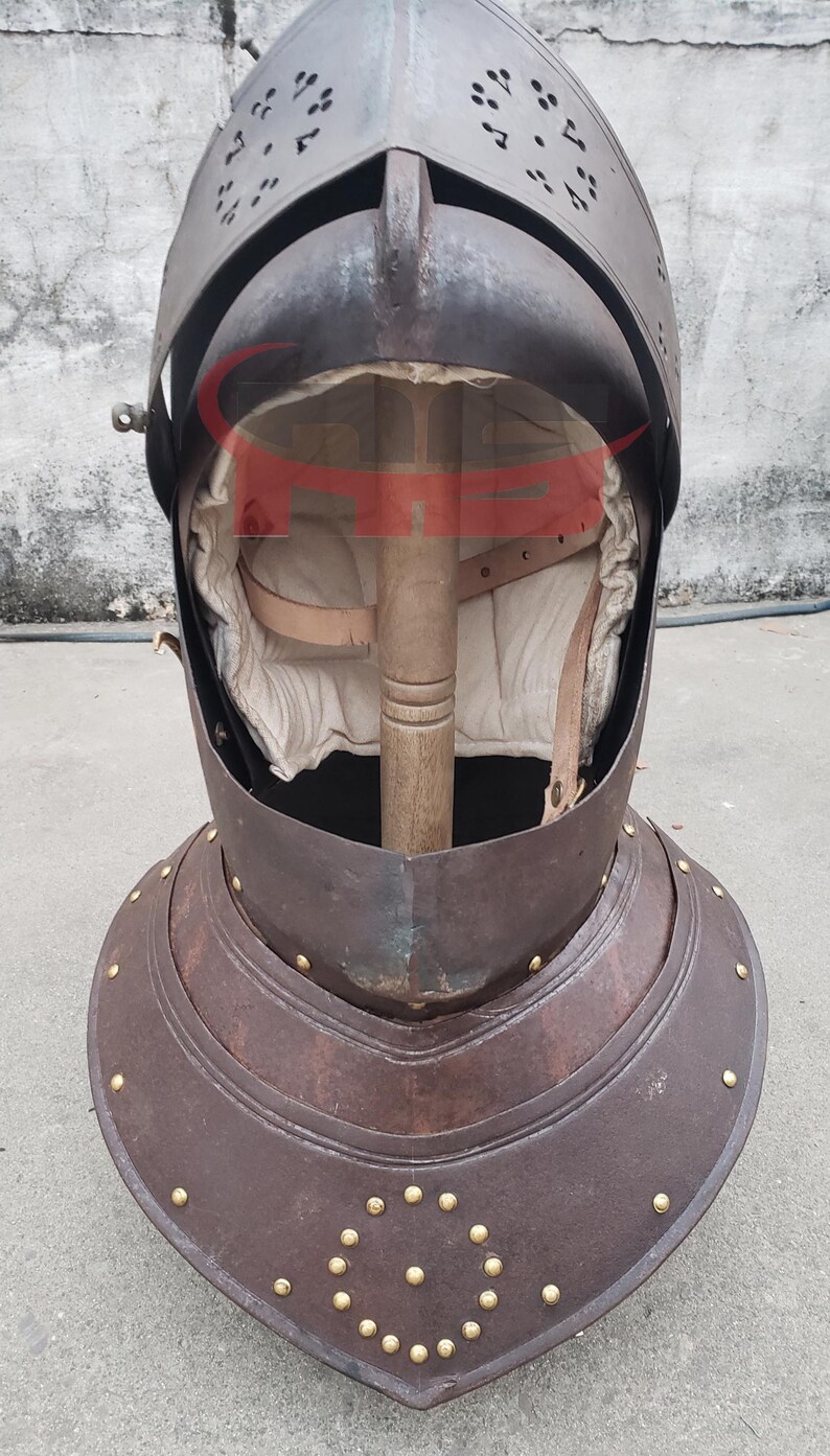 Puede incluir: Un casco medieval con un acabado marr&oacute;n oscuro y un borde de lat&oacute;n. El casco tiene una visera y una protecci&oacute;n para el cuello. El casco est&aacute; hecho de metal y tiene una superficie texturizada.