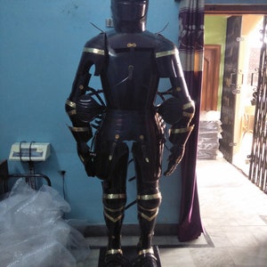 May include: A full suit of black armour with gold accents. The armour is standing on a black base. The armour is made of metal and has a helmet, breastplate, gauntlets, and greaves.
