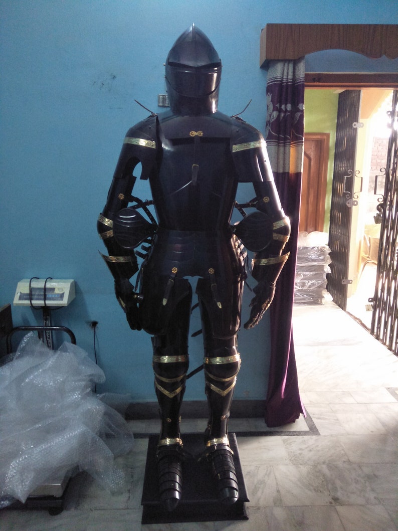 May include: A full suit of black armour with gold accents. The armour is standing on a black base. The armour is made of metal and has a helmet, breastplate, gauntlets, and greaves.