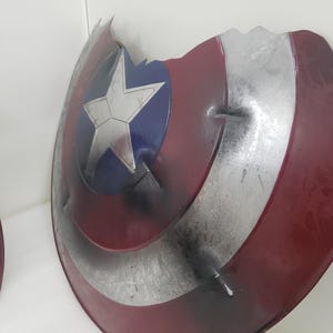 May include: A red, white, and blue Captain America shield with a silver star. The shield is cracked and damaged, with some of the paint chipped away.