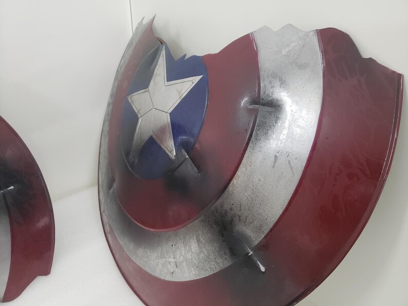 May include: A red, white, and blue Captain America shield with a silver star. The shield is cracked and damaged, with some of the paint chipped away.