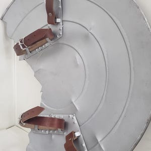 May include: A silver metal shield with a circular design and three brown leather straps. The shield is slightly dented and has a worn appearance.