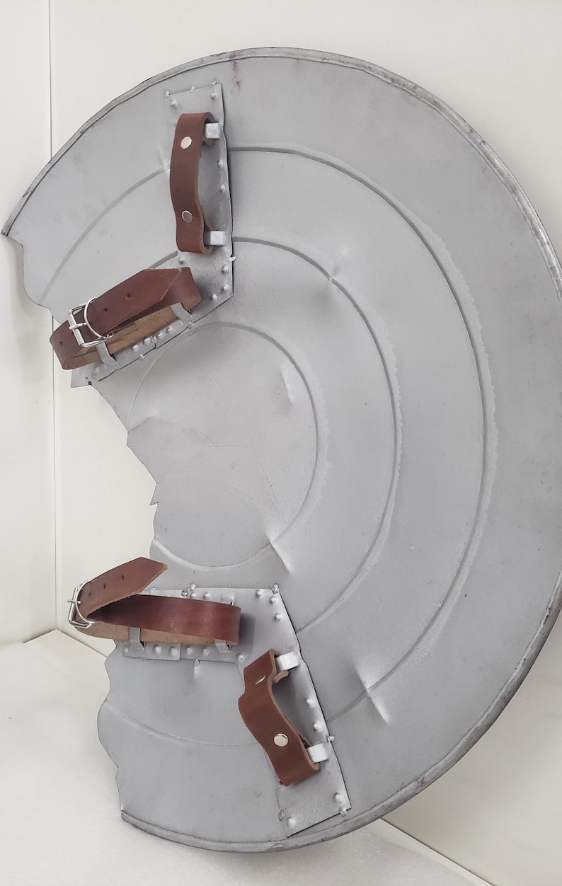 May include: A silver metal shield with a circular design and three brown leather straps. The shield is slightly dented and has a worn appearance.
