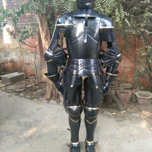 May include: Full body suit of black armour with gold accents. The armour includes a helmet, breastplate, gauntlets, and greaves. The armour is standing on a concrete surface.