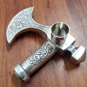 Puede incluir: Una pipa de plata con forma de hacha con un dise&ntilde;o ornamentado y una cazoleta extra&iacute;ble. La cazoleta es cil&iacute;ndrica y tiene un acabado liso y pulido.