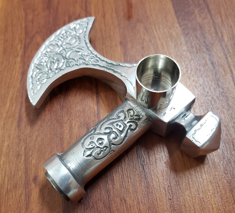 Puede incluir: Una pipa de plata con forma de hacha con un dise&ntilde;o ornamentado y una cazoleta extra&iacute;ble. La cazoleta es cil&iacute;ndrica y tiene un acabado liso y pulido.