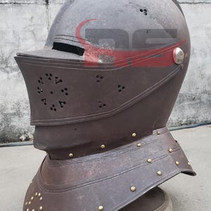 Puede incluir: Un casco de metal marr&oacute;n con visera y un dise&ntilde;o decorativo. El casco tiene un acabado en lat&oacute;n y est&aacute; sobre un soporte de madera.
