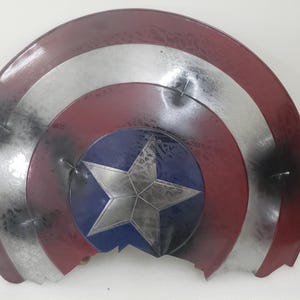 May include: A red, white, and blue Captain America shield with a large, silver star in the center. The shield is cracked and broken, with pieces missing.