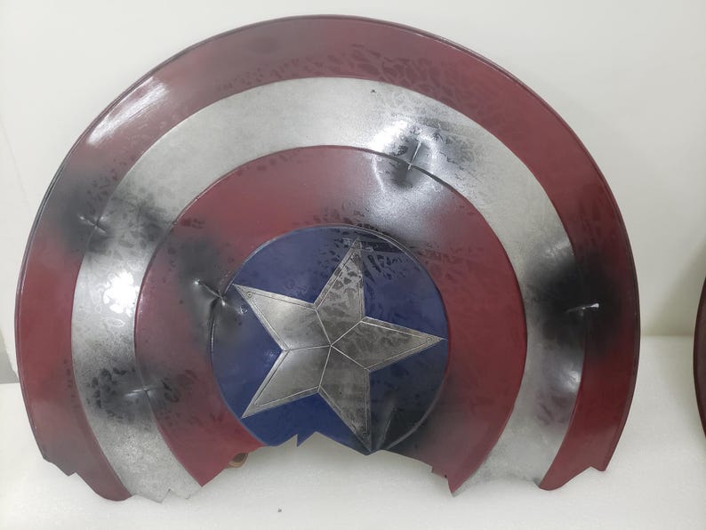 May include: A red, white, and blue Captain America shield with a large, silver star in the center. The shield is cracked and broken, with pieces missing.