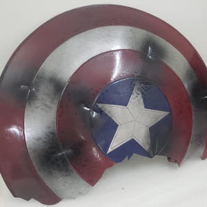 May include: A red, white, and blue Captain America shield with a star in the center. The shield is cracked and damaged, with black paint splatters on the surface.